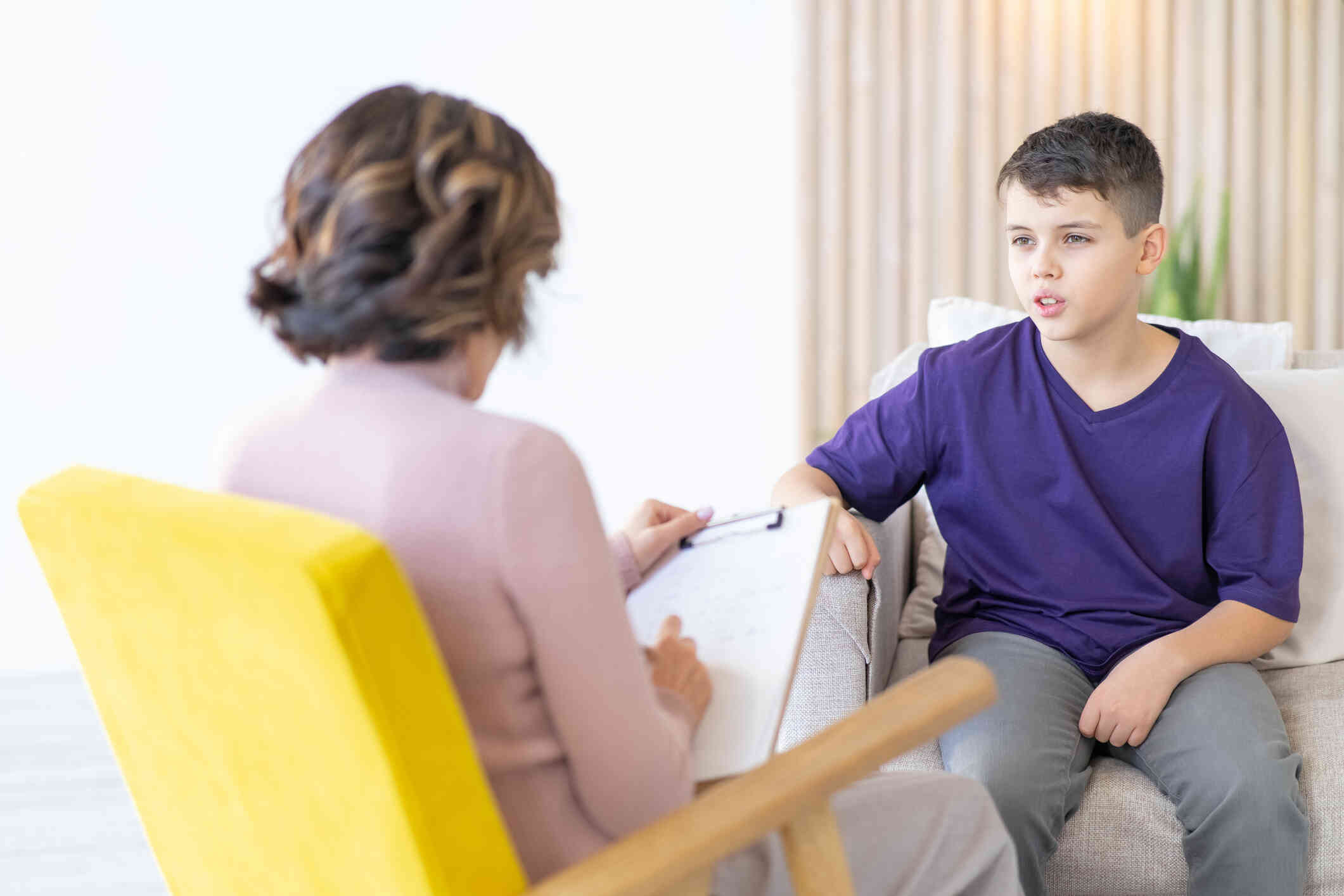 A boy in a purple shirt sits on a couch and talks to the female therapist sitting across from him during a therapy session.